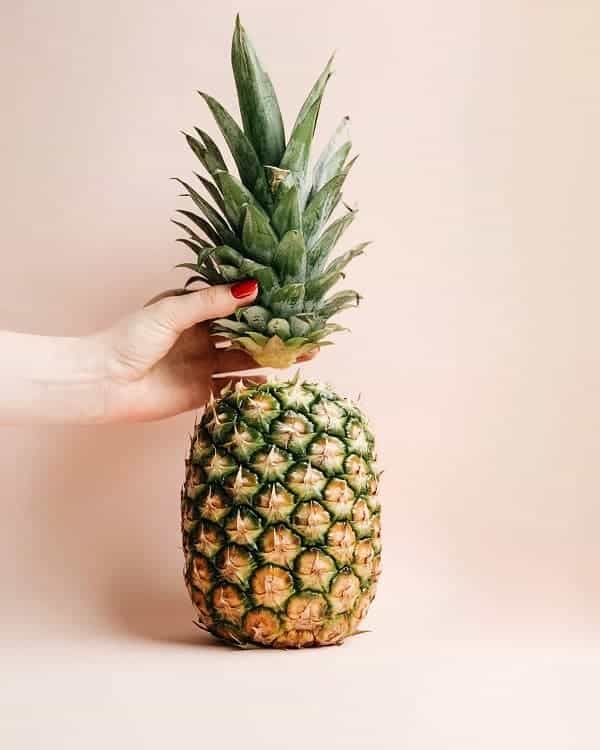 Hand removing a pineapple top to grow a pineapple houseplant, on light pink background.